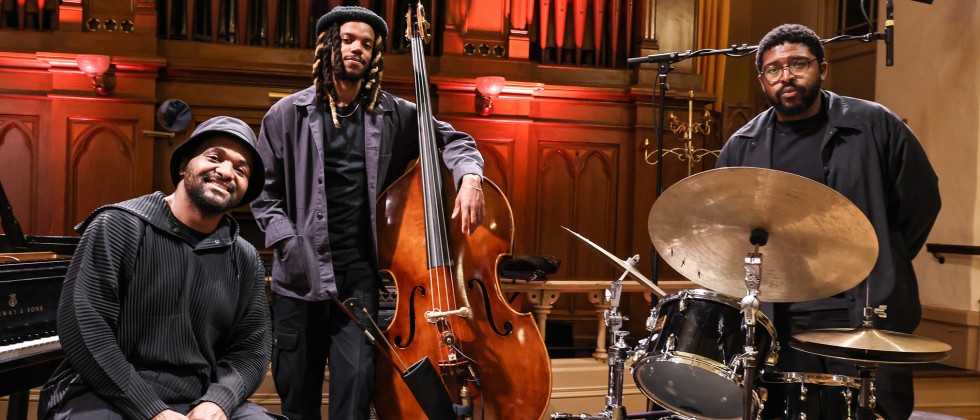 Three black men in black attire stand in front of organ pipes, one sits on a piano bench, the others pose with a bass and drum set. 