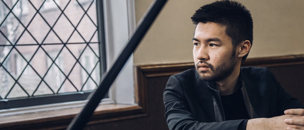 An Asian American man in a black jacket sits leaning on a grand piano, his face framed by the lid and the stick in a well-lit room. 
