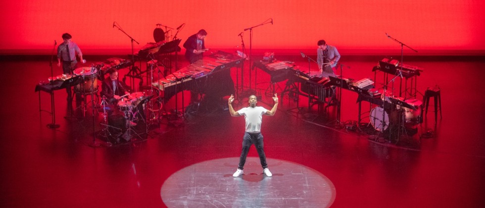 On a stage bathed in red, a single dancer stands in the center with his arms stretched upward, surrounded by mic'd percussion. 