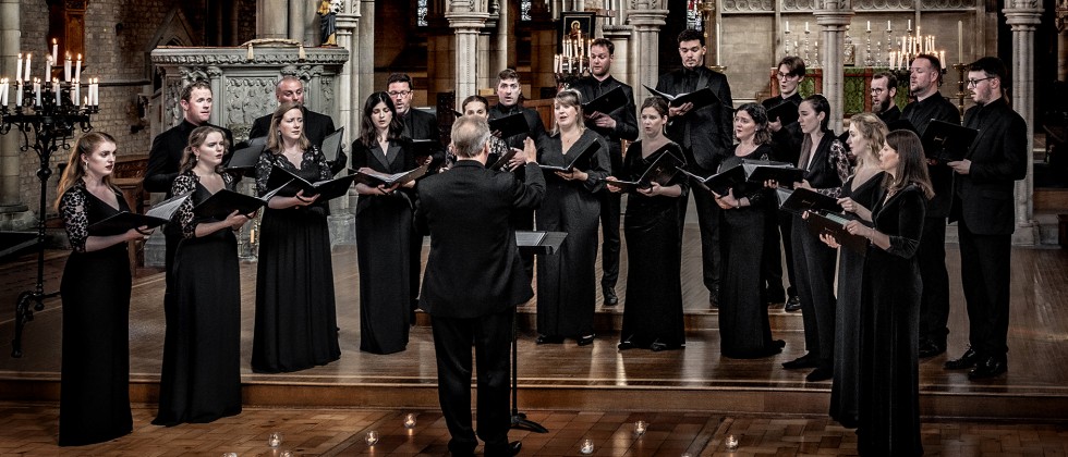 Tenebrae, A choir of twenty-one members stand holding binders of music, singing while conducted in an ornate church sanctuary-like room. 