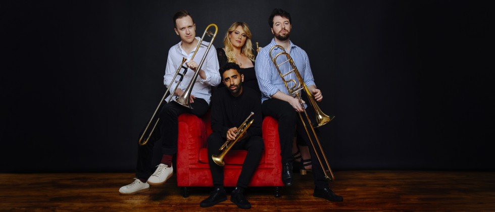 Two men sit on the arms of the chair holding trombones, one man sits centered in the chair holding a trumpet, behind him a woman with long blond hair in a black dress leans into the frame. 