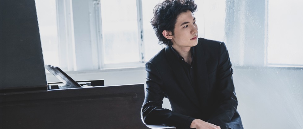 In front of a glassy window, a man with dark, curly hair sits in a black jacket leaning on the lid of the keys on a grand piano. 