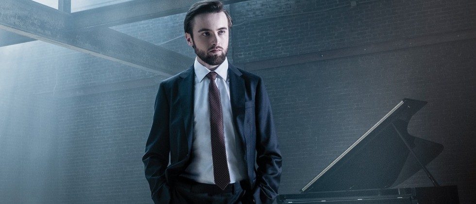 A young bearded white man, under cool light in a blue suit in an industrial building, stands with his hands in his pockets. A grand piano is in the background.