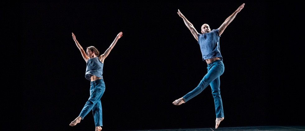 Two Trisha Brown Dance Company members leap with arms straight and pointing upwards in a V with one leg pointing down and one pointing a cross their body. 