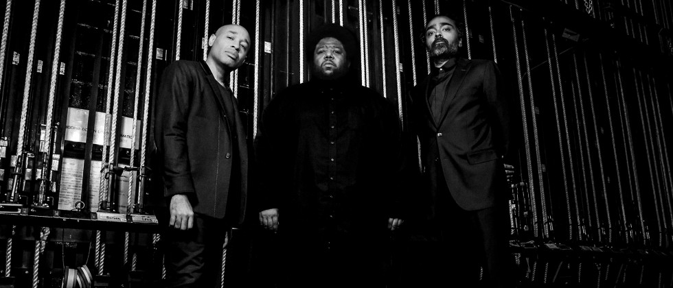 A black and white photo of three stoic men standing in front of backstage rigging. 
