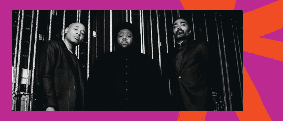 A black and white photo of three stoic men standing in front of backstage rigging. 