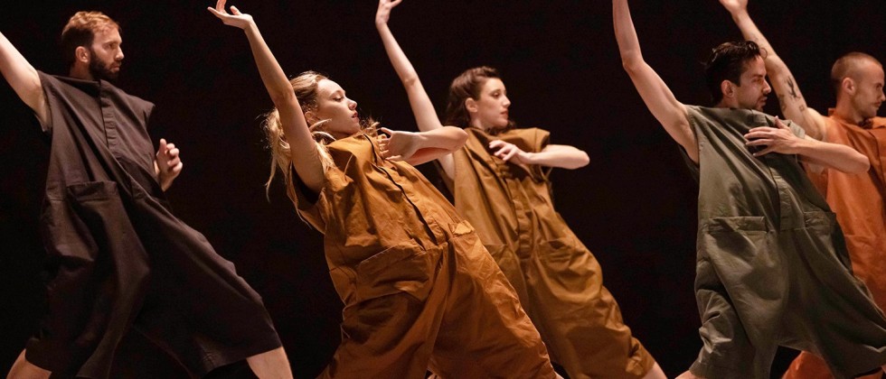 a dance company, men and women wearing baggy rompers in earthy colors. their feet are apart, and they lean back, each with an arm flung above their head.