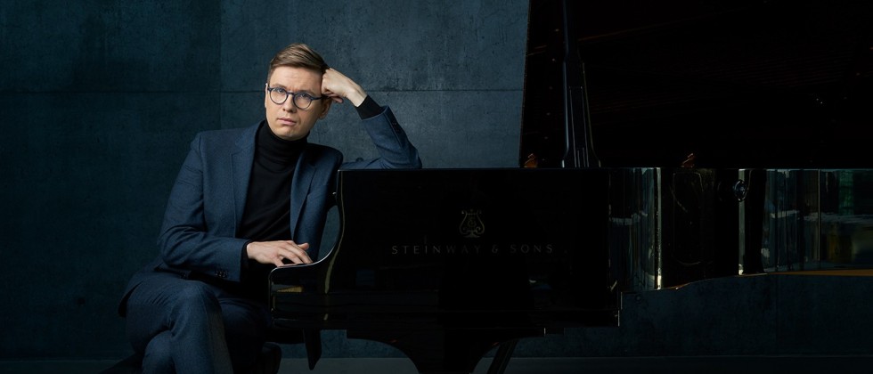 A white man with round glasses in a blue suit in front of a blue background rests an elbow on the top of a piano, his fist at his temple and his other hand idly playing the keys without looking.