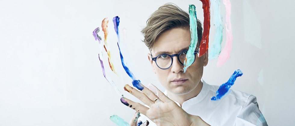 Vikingur Olafsson, A white man with small round glasses stares intensely ahead while using finger paint on a plexiglass panel in front of his face. 