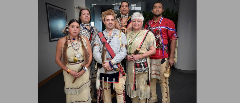 A mixed group of six Indigenous performers in traditional dress stand together with gentle smiles in a white hallway. 