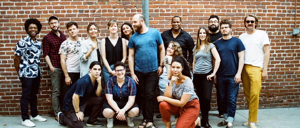 A group of 16 people in casual outfits stand and kneel outdoors in front of a red brick wall.