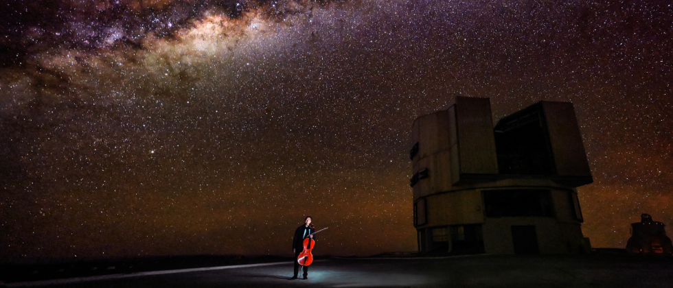 Yo-Yo Ma, under a small white spotlight in the Atacama Desert. The galaxy spans the dark sky above him, and a large rock outcrop looms at his left, a dark space in the bright sky..