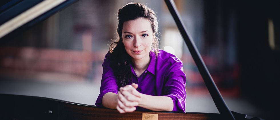 A white woman with pulled-back long brown hair wearing a bright purple shirt as she leans over the keys of a grand piano. 