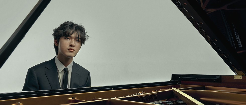 A young, South Korean man sits at the piano, his face framed by the stick of the lid, staring solemnly ahead in a dark suit. 