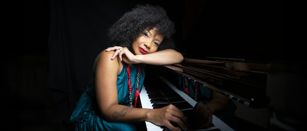 A Cuban woman with dark curly hair leans her head on her arm on the piano while another hand lays on the piano keys. 