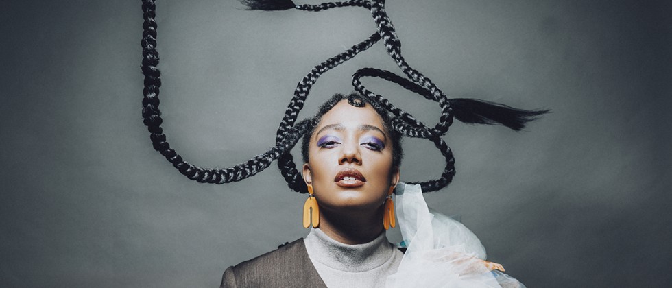 A black woman in a black, gray, and blue top with blue/white tulle stands looking up, while four braids twist into the open space above her head. 