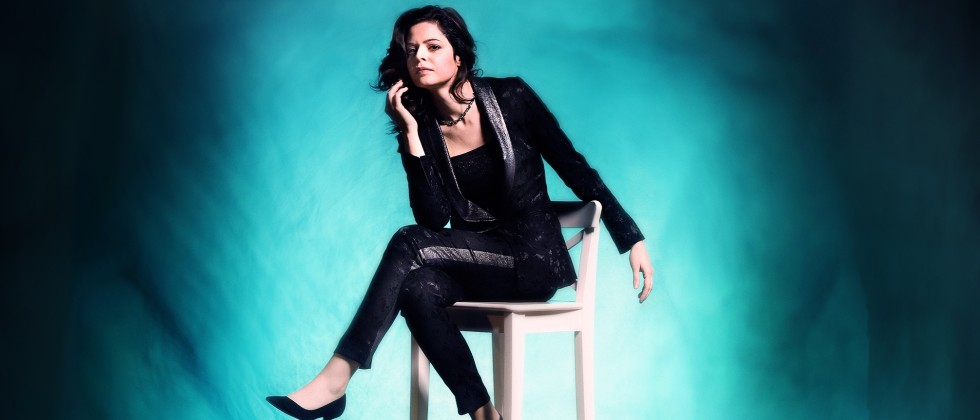 A woman in a black leather, textured outfit sits on a tall chair with her legs crossed before a glowing turquoise background. 