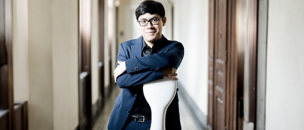 A young man with dark hair and square glasses in a blue suit stands leaning on a hard, white cello case. 