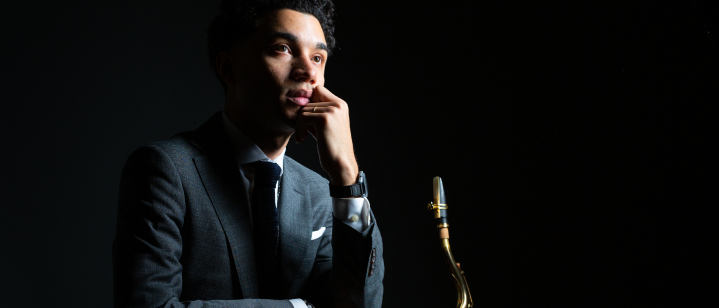 A Puerto Rican man with dark, curly hair sits holding a tenor saxophone while leaning his chin on his left arm. 