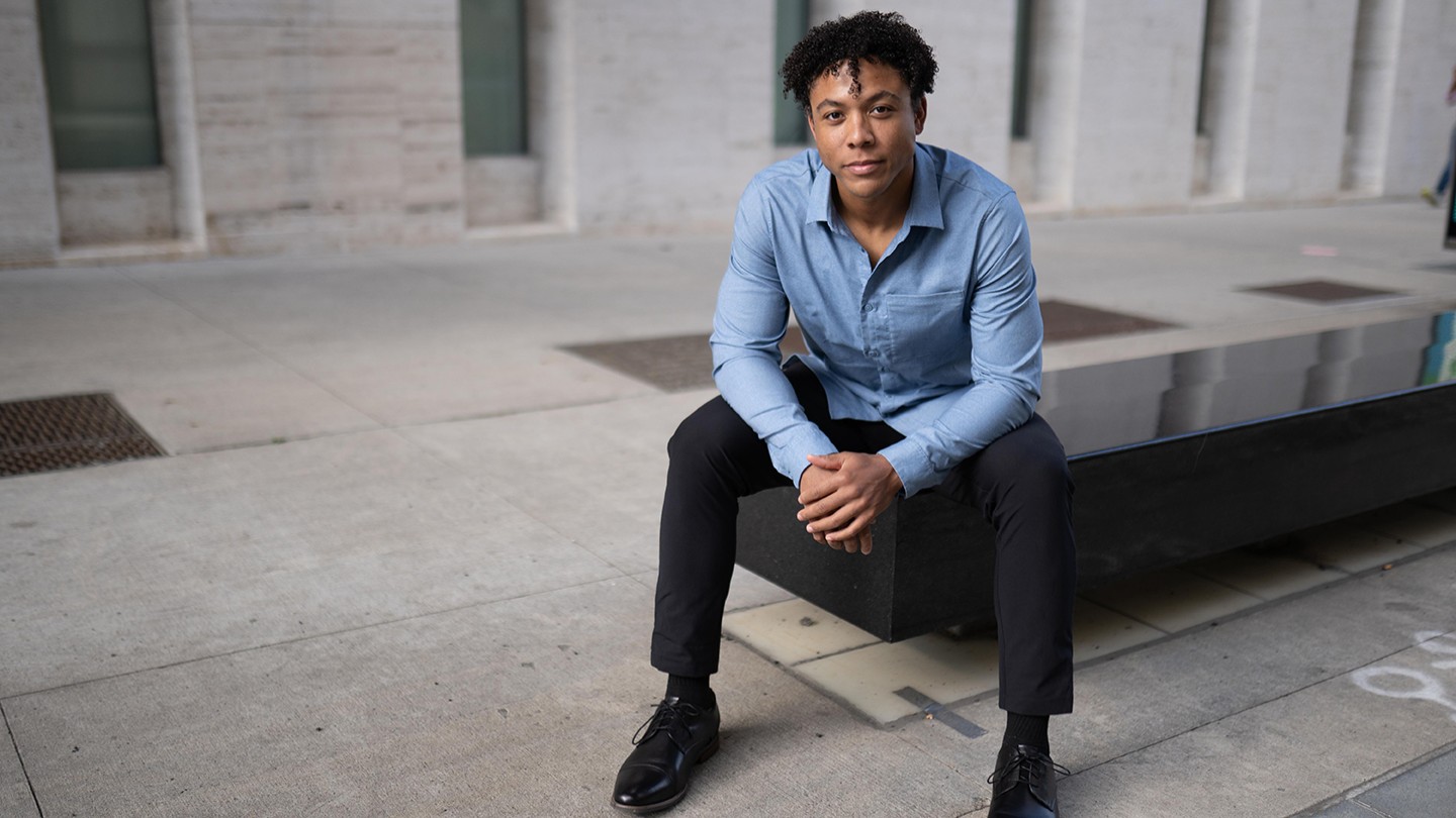 A young Black sits on the edge of a stone bench, his elbows resting on his knees and his hands crossed. He wears a soft blue collared shirt..