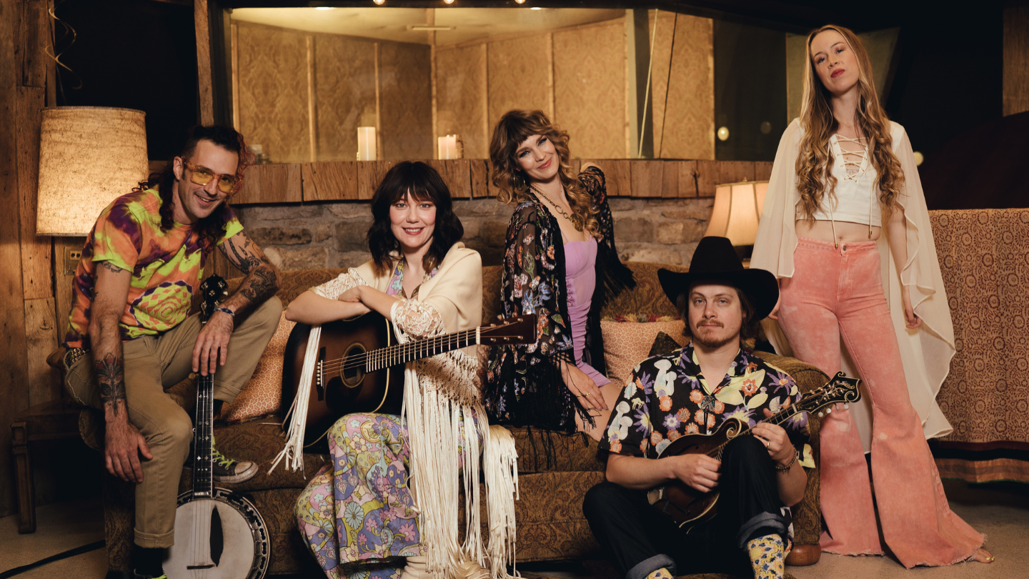 In a wood-paneled room, five young bluegrass musicians smile, as they lounge with their instruments on a brown velour couch. Their style is eclectic, with a retro 1960s-70s flair