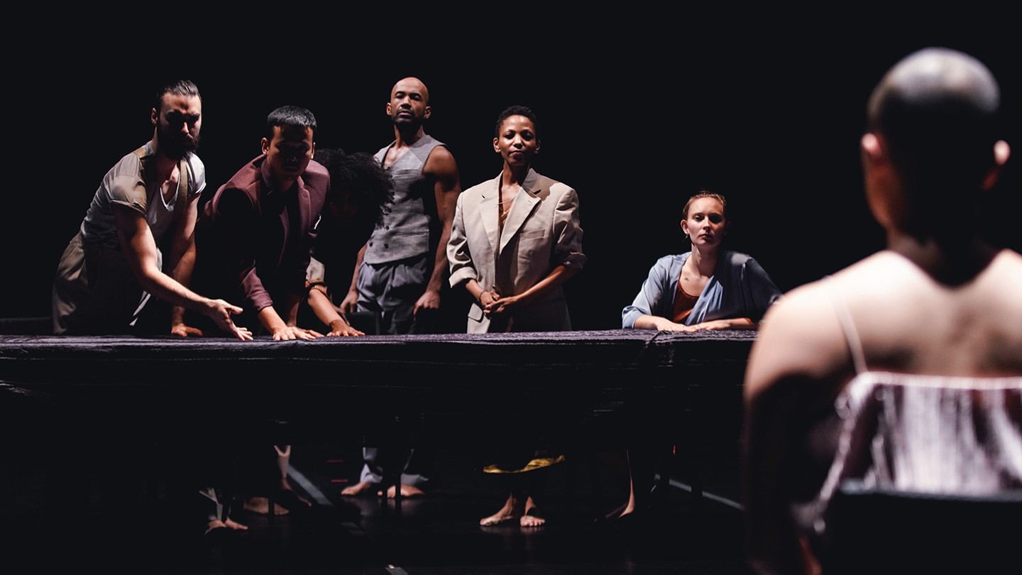 a diverse group of five men and women lean over a long table, as if challenging a person whose back is turned to us