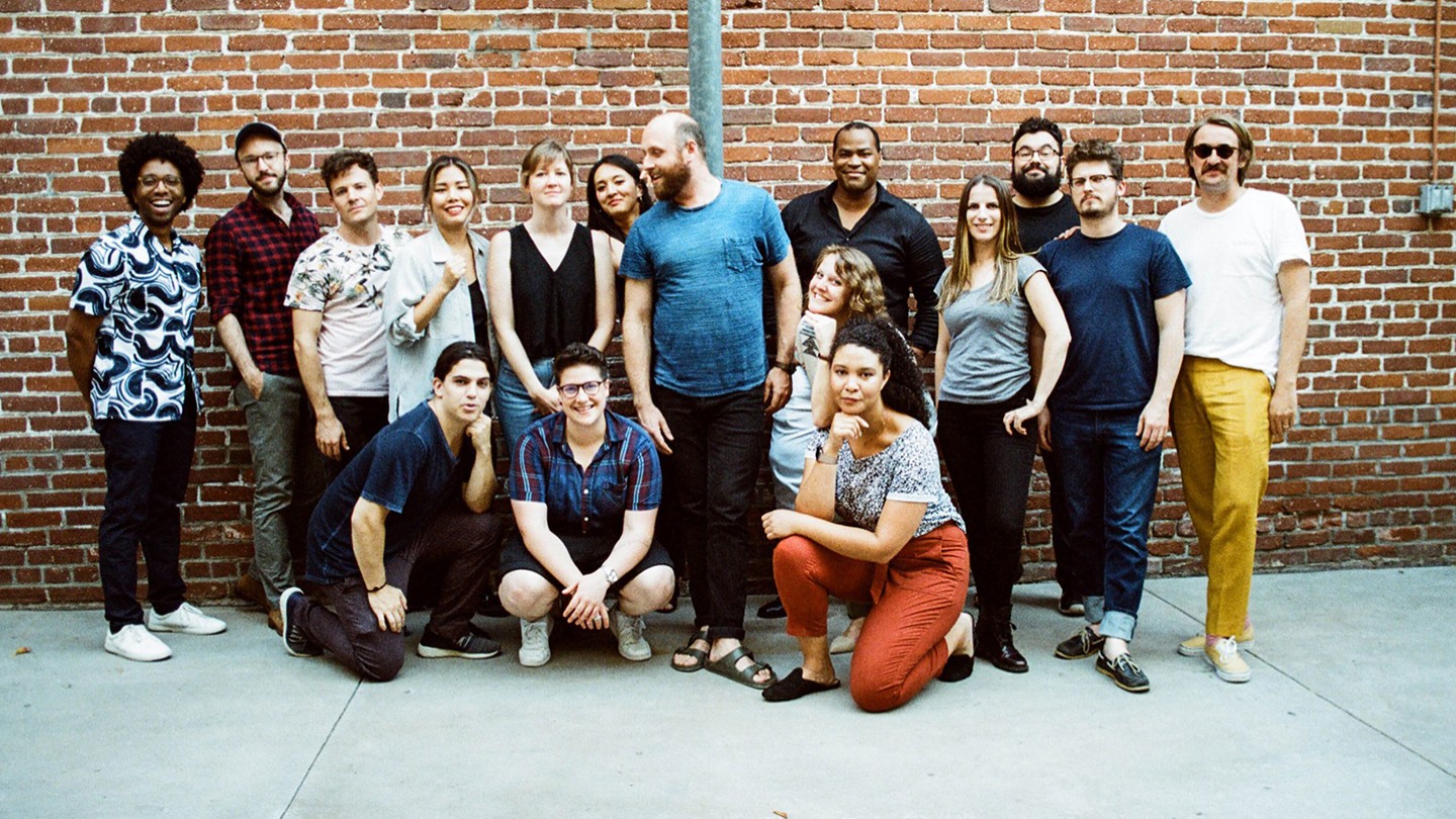 A group of 16 people in casual outfits stand and kneel outdoors in front of a red brick wall.