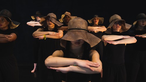dancers, each with their forearms stacked in front of them at shoulder level, stand in a phalanx wearing floppy woven hats