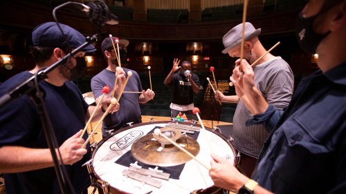 gathered around a bass drum turned on its side with percussion instruments on top, four members of a percussion duo and a beatboxer rehearse