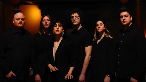 Six people in a shadowy room stand in a line wearing concert black and staring forward with serious expressions. 