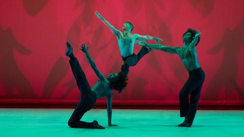 three male dancers, all shirtless in black pants and dramatically lit in red and green, with their arms and legs at extreme angles in the middle of an energetic dance.