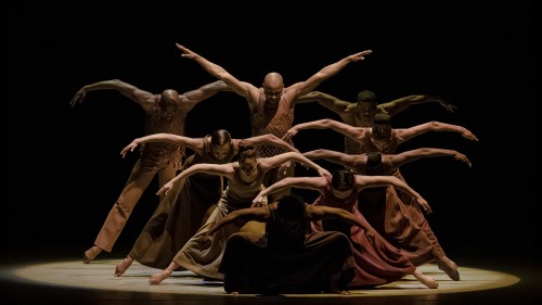 Several dancers are arranged in a series of lines, each dancer standing taller than the one in front, with arms arched beside them. 