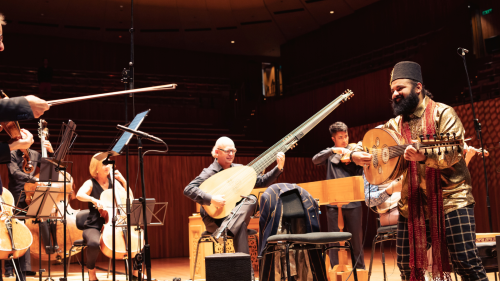 white man in a black suit conducts a chamber orchestra, featuring a smiling Egyptian Australian man with a curly mustache, a thick beard, and a cylindrical maroon hat