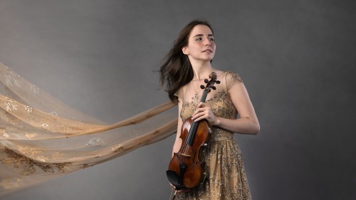Before a gray photo backdrop, a young Armenian woman in a tan dress holds a violin. An embroidered scarf billows behind her.