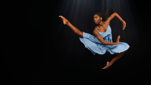 a dark skilled black woman leaps beofre a black background, one leg extended, one tucked under. Her arms, swept to her side, make a circle.