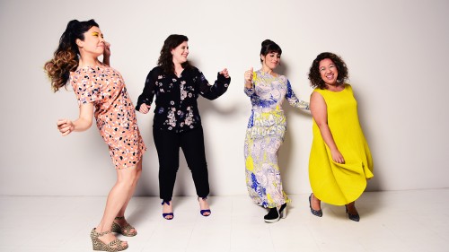 Aizuri Quartet, four women in patterned and solid clothing, stand in front of a white wall dancing and smiling. 