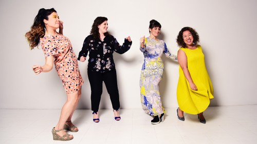Aizuri Quartet, four women in patterned and solid clothing, stand in front of a white wall dancing and smiling. 
