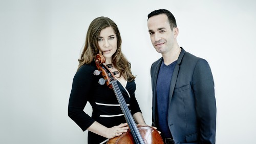 A white man and woman with long brown hair stand close together in front of a white background; the man wears a blue/gray suit, and the woman wears a black dress with white highlights while holding her cello. 
