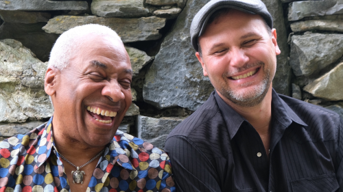 Two men sit laughing in front of a stone wall; on the left a Black man with short white hair wears a colorful polka-dotted shirt with a silver turtle necklace; on the right a white man with a trimmed goatee wears a black striped shirt and a flat gray cap.