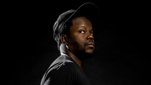 Black man, Ambrose Akinmusire in a black shirt and hat in front of a black background. 
