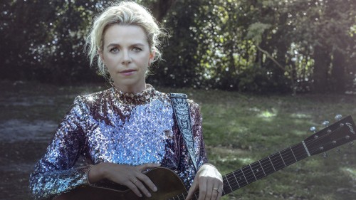 A white woman with blond hair and a metallic sequins dress, and an acoustic guitar stands in front of a tree-lined field.