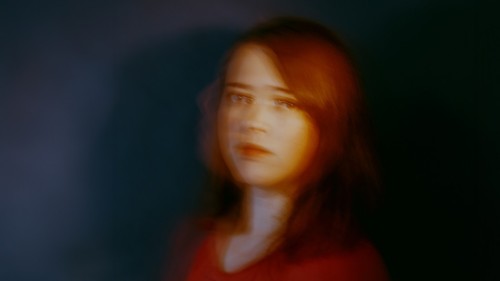 A white woman with shoulder length red hair and red shirt is pictured in front of a dark background in an intentionally blurry portrait. 