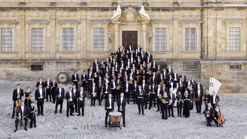 The members of the bamberg symphony, arrayed with their instruments on a cobblestone plaza in front of the steps of a yellow stone building 