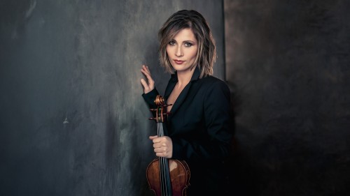 A white woman with shoulder length brown hair leans against a gray painted wall holding her violin while wearing a dark suit jacket. 