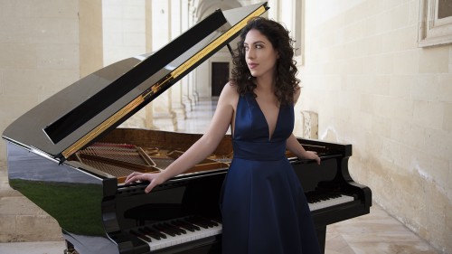 Beatrice Rana, a woman in a blue dress stands with her arms extended over a piano. 
