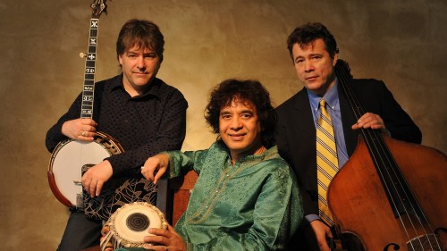 Bela Fleck, a white man wearing a black shirt holding a banjo, Zakir Hussain, a man in a green shirt sitting down, and Edgar Meyer, a white man in a blazer with a yellow tie holding a bass, in front of a brown background. 