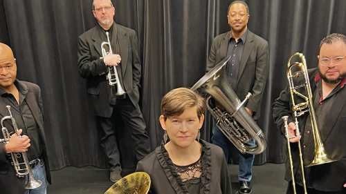 In a black box space, a mixed group of four men stand in an arch with a white woman standing in front. They all wear black jackets while holding brass instruments and stare intensely forward. 