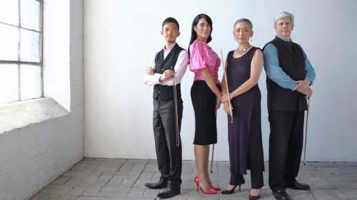Four performers of different races and ages stand in a row, back-to-back, in front of a white background and stone floor. They each are holding the bows to their string instruments and smile stoically at the camera. 