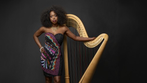 A Black woman poses with a golden harp in front of a dark background in a short, strapless dress with a multi-colored geometric pattern. 