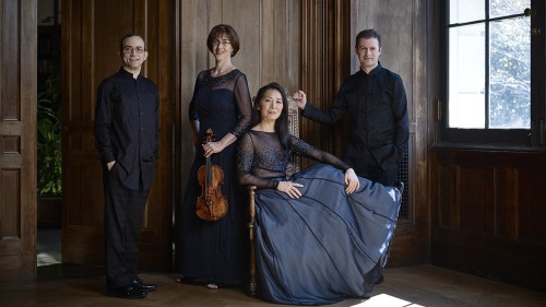 the four members of a string quartet, two men flanking two women, inside an ancient mansion. One of the women holds a violin.